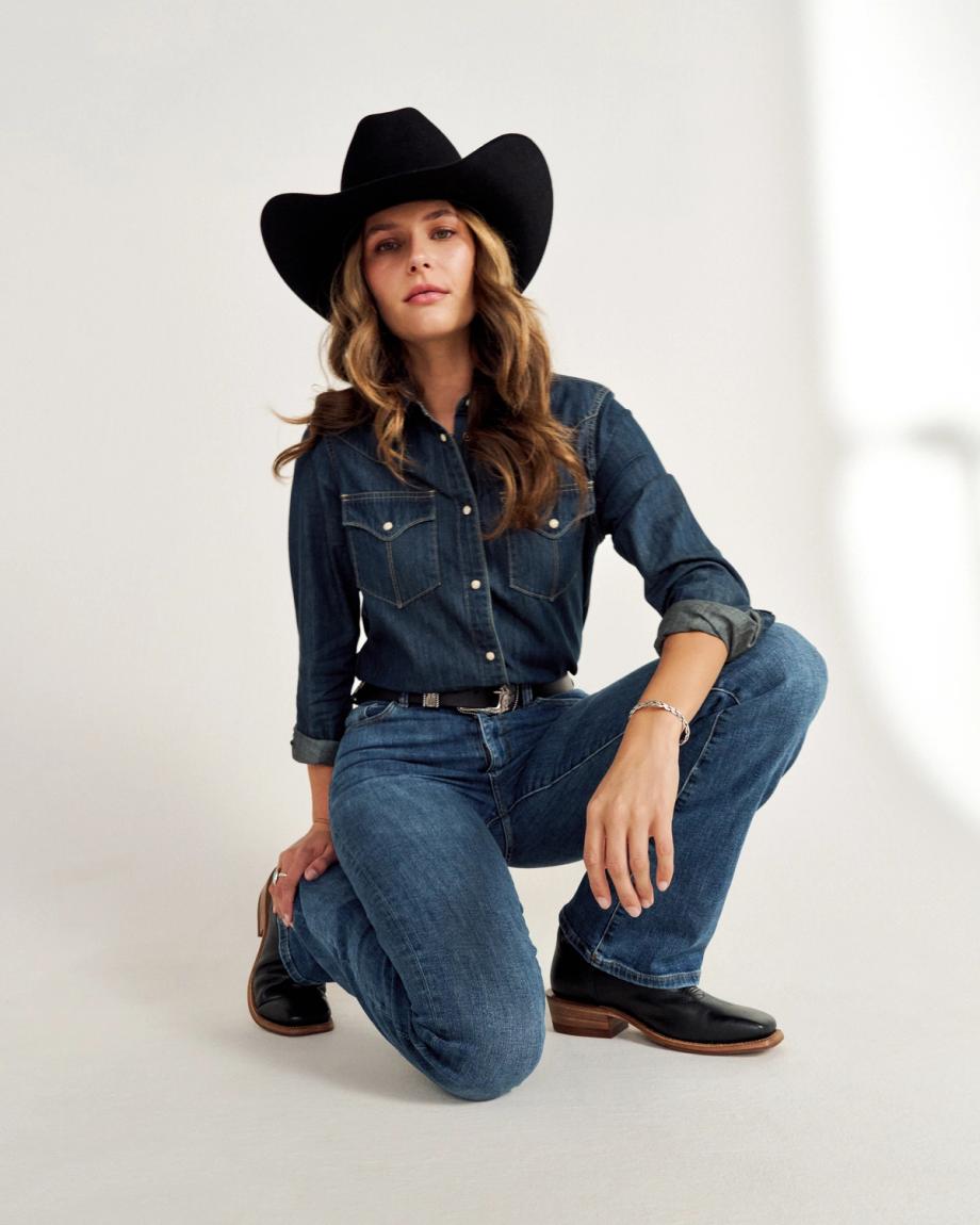 A person in a cowboy hat kneels in a denim outfit against a simple backdrop.