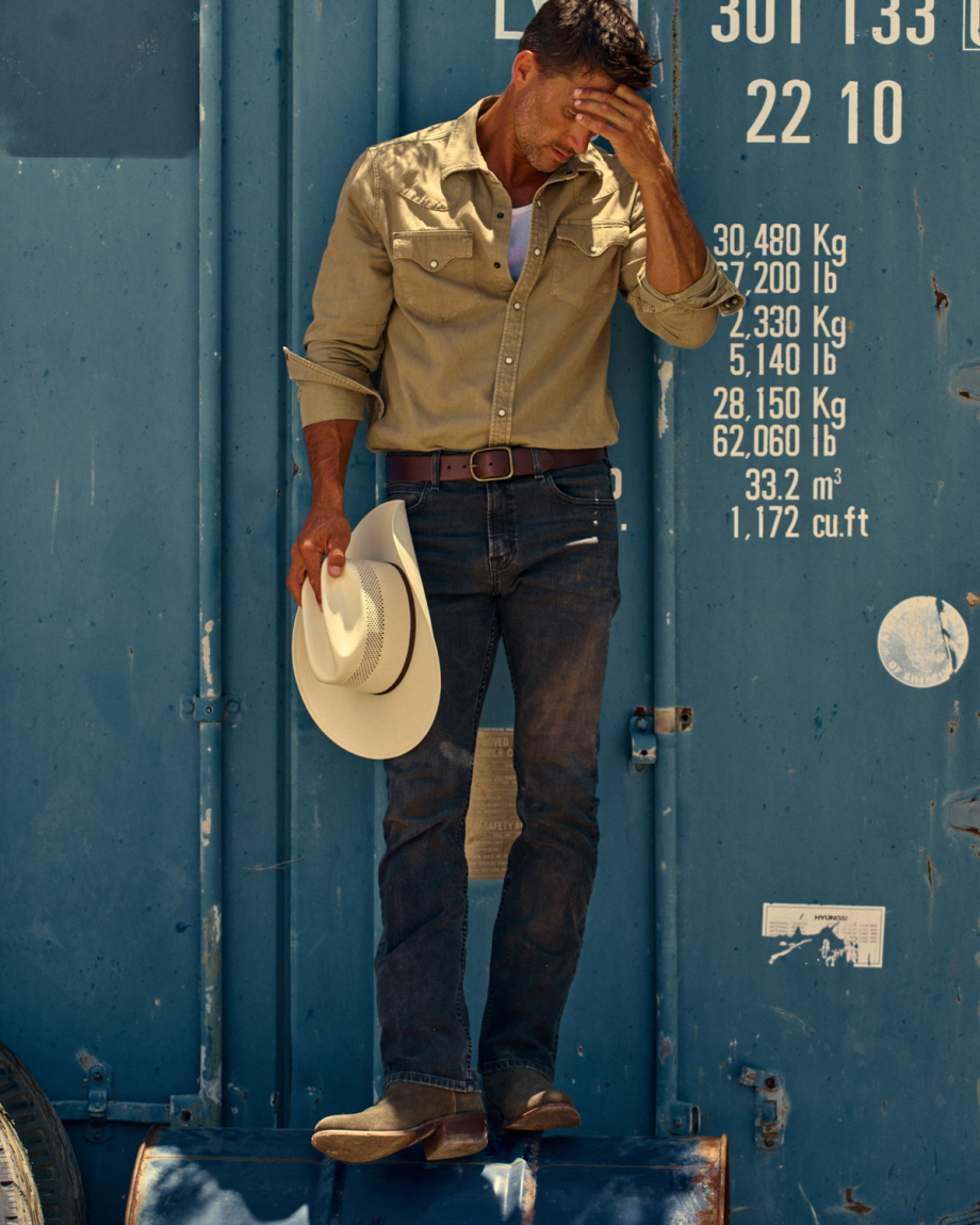A man in a tan shirt and jeans stands in front of a blue shipping container, holding a white cowboy hat and covering part of his face with his hand.