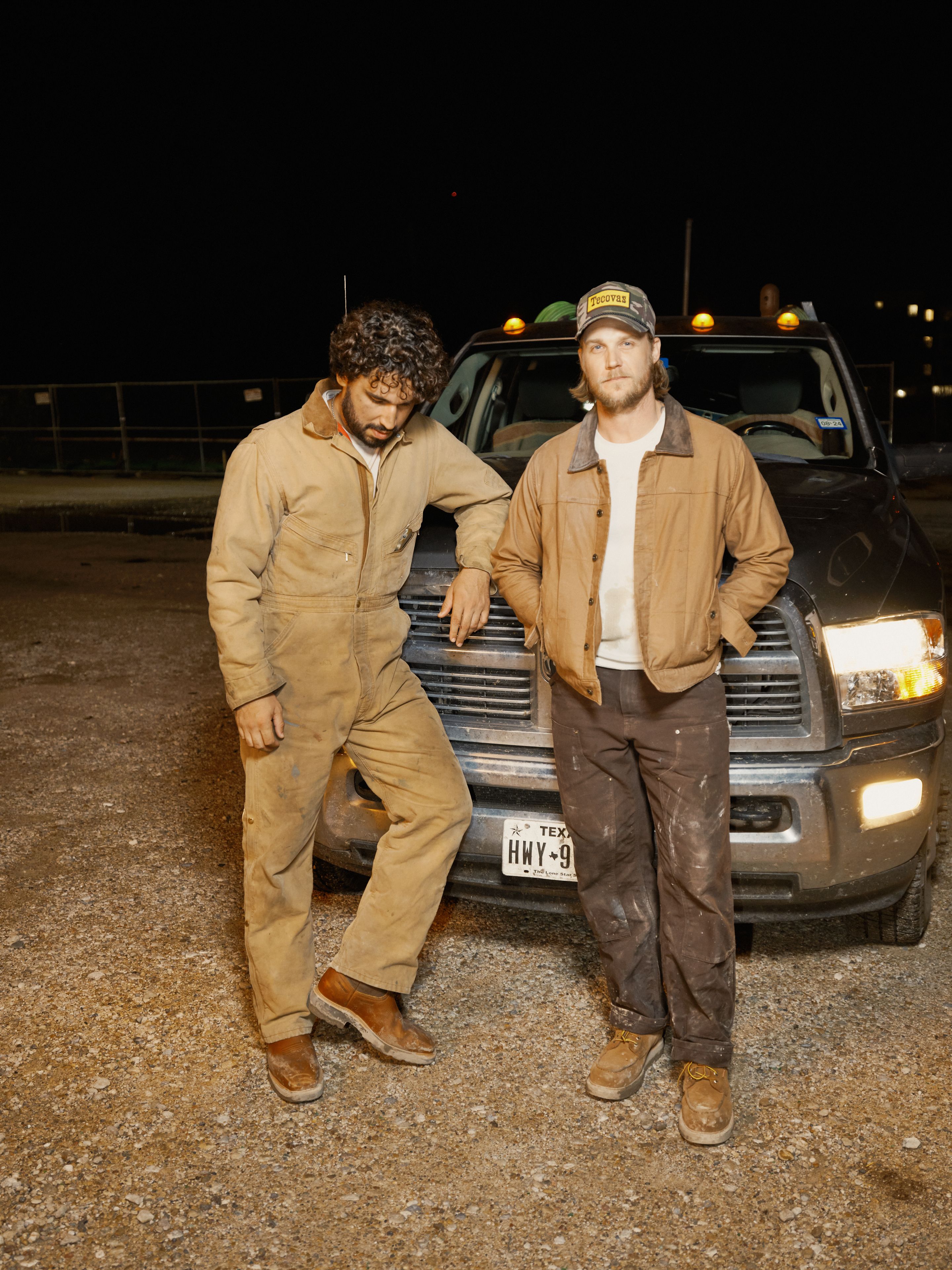 Two men wearing work clothes stand in front of a truck at night. One leans against the vehicle, while the other stands with arms crossed.
