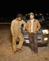 Two men wearing work clothes stand in front of a truck at night. One leans against the vehicle, while the other stands with arms crossed.