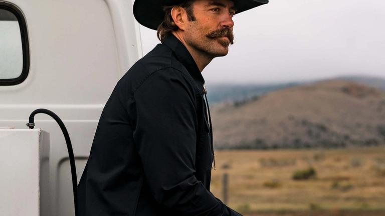 Image of a man in a cowboy hat sitting on the back of a pickup truck in the mountains.