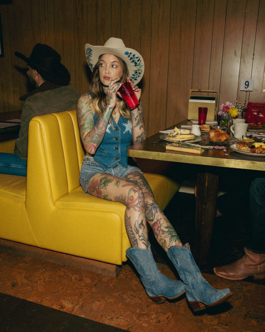 Person with tattoos sits on a yellow booth seat, wearing a cowboy hat, denim outfit, and boots, sipping from a red cup. Other patrons are in the background. Plates of food are on the table.
