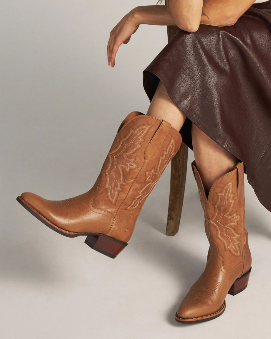 The Britt in Latte Goat. Person sitting in a chair wearing a dark brown skirt and tan cowboy boots with decorative stitching, sipping a creamy Latte Goat coffee.