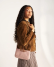 A woman with long curly hair wearing a brown fringed suede jacket and a checkered skirt, holding a light pink shoulder bag, stands against a plain background.