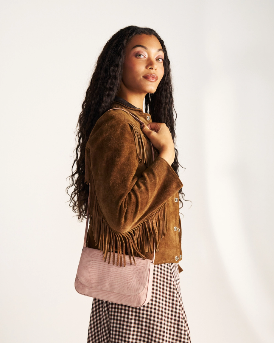 A woman with long curly hair wearing a brown fringed suede jacket and a checkered skirt, holding a light pink shoulder bag, stands against a plain background.