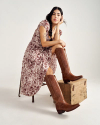 A woman in a patterned dress sits on a chair with her hand on her face, wearing brown knee-high boots.