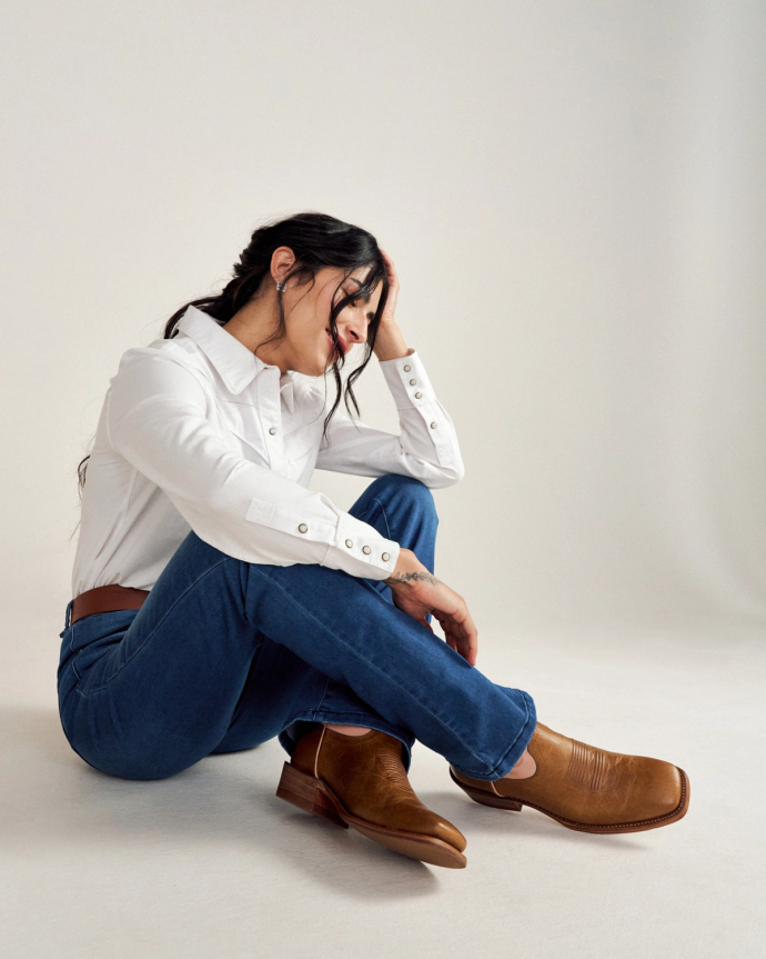 A person wearing a white shirt and jeans sits on the floor, resting their head in one hand, with brown boots visible.