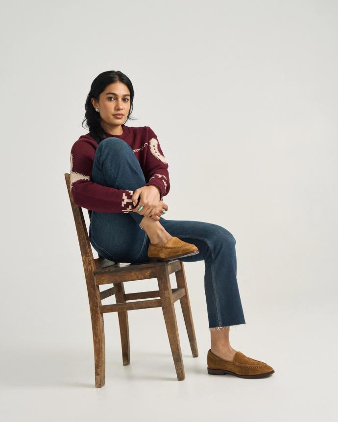 A woman sits sideways on a wooden chair, wearing a red patterned sweater, blue jeans, and brown loafers against a plain background.