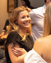 A woman with long blonde hair smiles and talks to another person at an indoor social gathering.