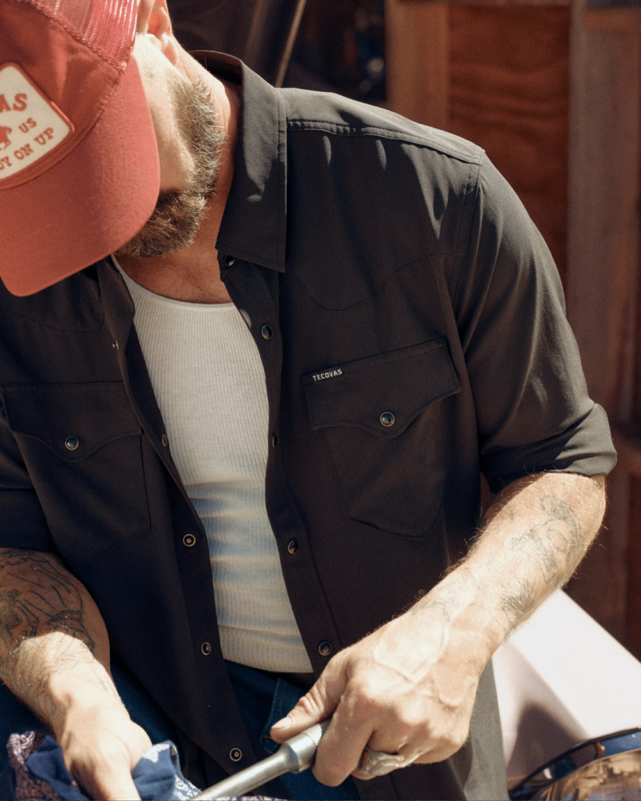 Man wearing a red cap, dark button-up shirt, and white undershirt is looking down while cleaning or working with his hands outdoors.