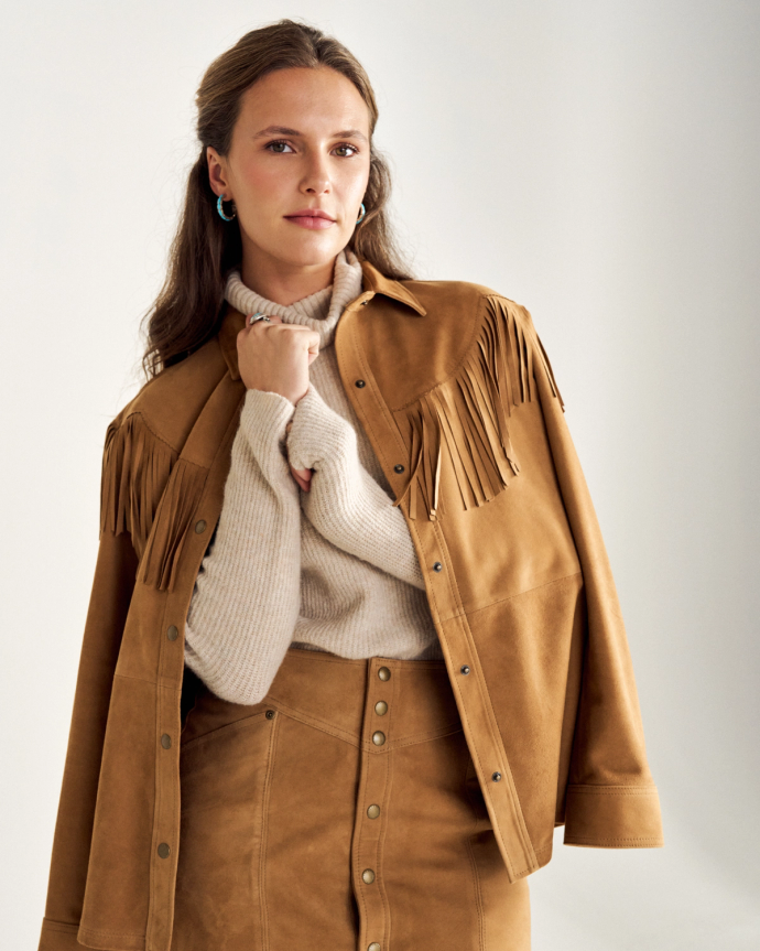 A person wearing a tan fringed jacket and skirt over a cream sweater poses against a plain backdrop.