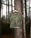 A camouflage-patterned T-shirt on a hanger is displayed against the trunk of a tree in a wooded area.