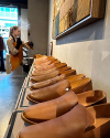 A row of brown leather slip-on shoes displayed on a shelf in a store, with a person in the background using a steamer.