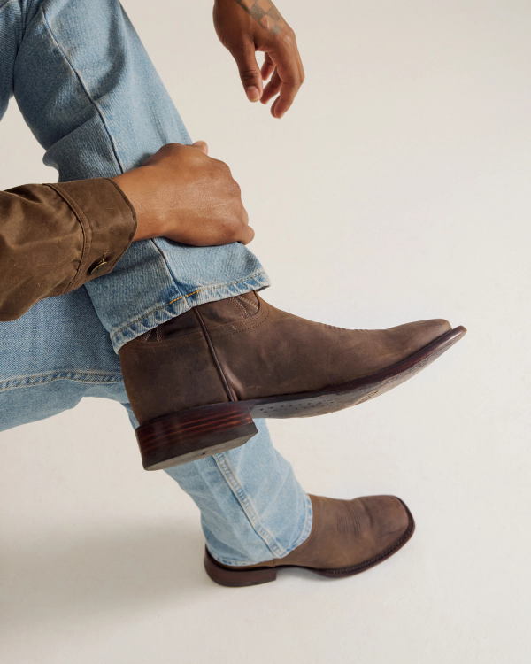 Person wearing light blue jeans, a brown shirt, and brown leather boots, sitting with one leg crossed over the other against a plain light background.