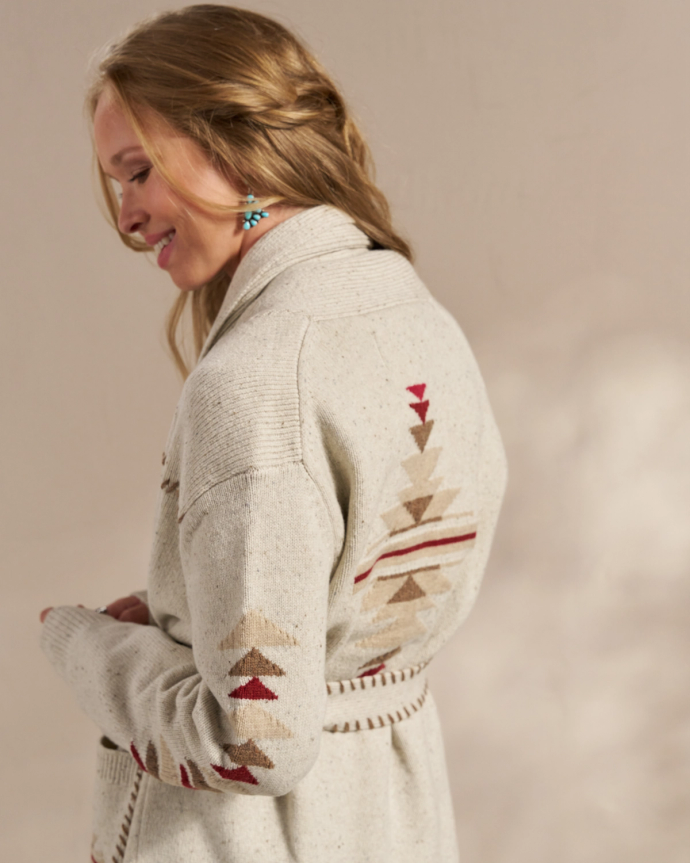 A person with long hair is wearing a light-colored, patterned cardigan and turquoise earrings, facing slightly to the right and smiling.