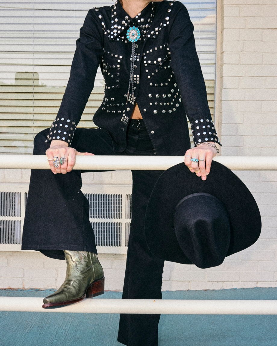 The Sadie in Olive Cowhide. Person in a studded black suit with turquoise jewelry sits on a railing, holding a black cowboy hat and wearing green Olive Cowhide boots, in front of a white brick wall and window.