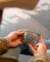 A person wearing jeans, boots, and a cowboy hat holds an intricately engraved silver wallet while seated on a wooden floor.
