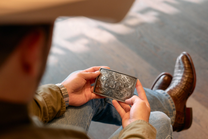 A person wearing jeans, boots, and a cowboy hat holds an intricately engraved silver wallet while seated on a wooden floor.