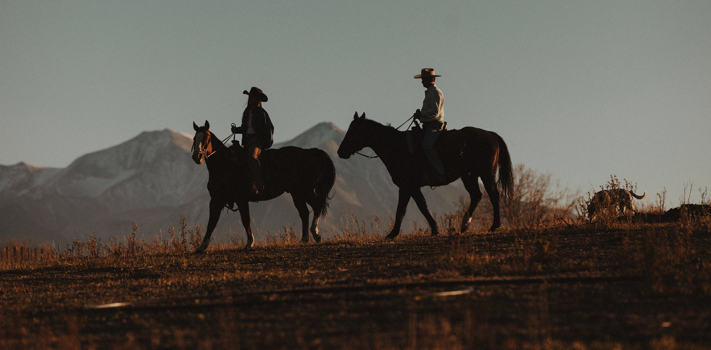 Two people in cowboy hats ride horses across a ridge with mountains in the background.