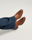 A person wearing blue jeans and The Dean Ostrich pecan cowboy boots sits with their legs crossed against a plain background.