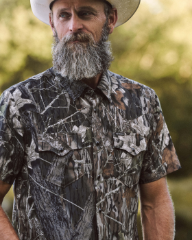 Man wearing a camouflage short-sleeve shirt and white cowboy hat stands outdoors, with trees blurred in the background.