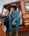 Person sitting on the edge of an open truck door, wearing a denim jacket, hat, jeans, and boots, in a rural setting.