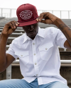 Man in a white button-up shirt adjusts a burgundy baseball cap with white text while seated on outdoor bleachers.