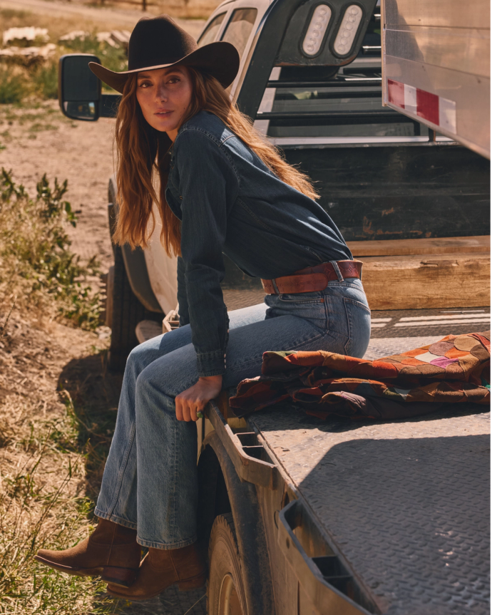 A person wearing a cowboy hat and denim outfit sits on the back of a truck parked on a dirt path.