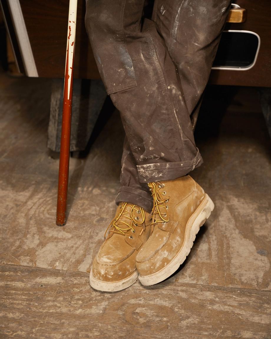 Person wearing muddy work boots and dark pants, leaning against a surface with a red-handled tool beside them.