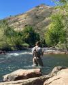 A person in fishing gear casts a line in a rocky river with a hill in the background under a clear sky.