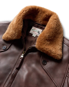 A brown leather jacket with a light brown fur collar, featuring a front zip and button closure. The jacket has a label with text sewn on the inner collar.