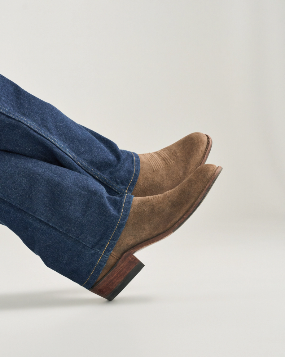 The Johnny in Gravel Suede. A person wearing blue jeans and Gravel Suede cowboy boots with pointed toes, viewed from the side against a plain background.