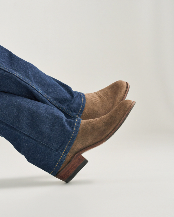 The Johnny in Gravel Suede. A person wearing blue jeans and Gravel Suede cowboy boots with pointed toes, viewed from the side against a plain background.