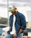 Man outside wearing the Tecovas Five-panel mid profile trucker hat in the color cream/olive. 