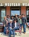 A group of people in cowboy hats pose in front of a store named "Howell Western Wear" with some seated on a bench and others standing behind them.
