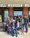 A group of people in cowboy hats pose in front of a store named "Howell Western Wear" with some seated on a bench and others standing behind them.