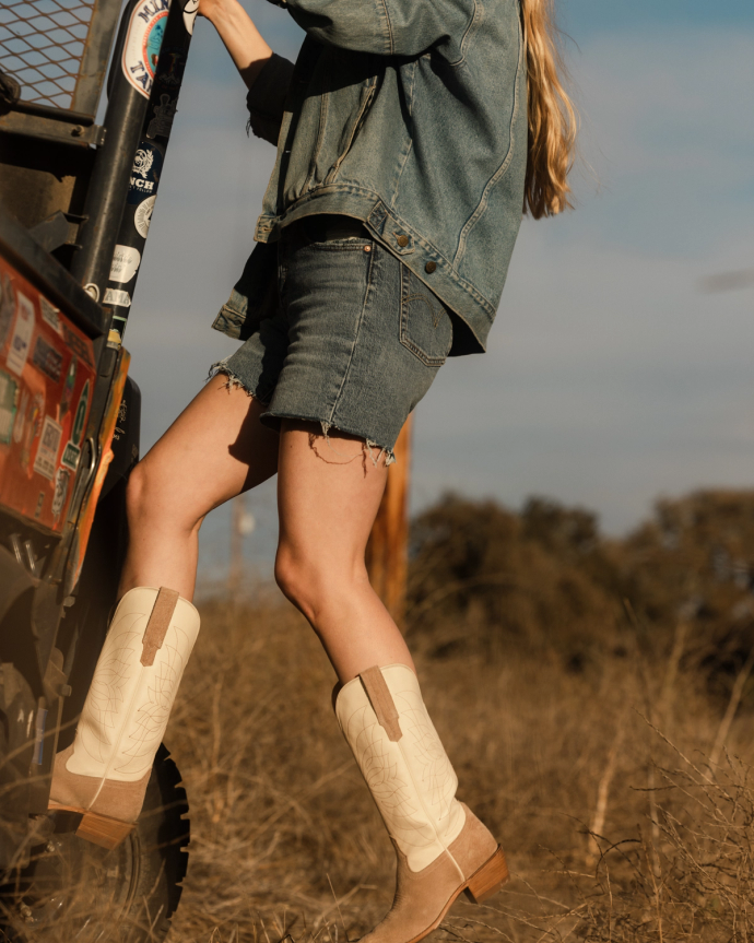 Person wearing denim jacket and skirt with cowboy boots, stepping onto a vehicle in a grassy field.