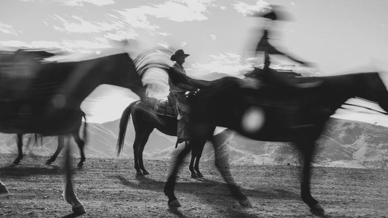 Black and white photo of blurred riders on horseback, with mountains in the background and partly cloudy sky above.