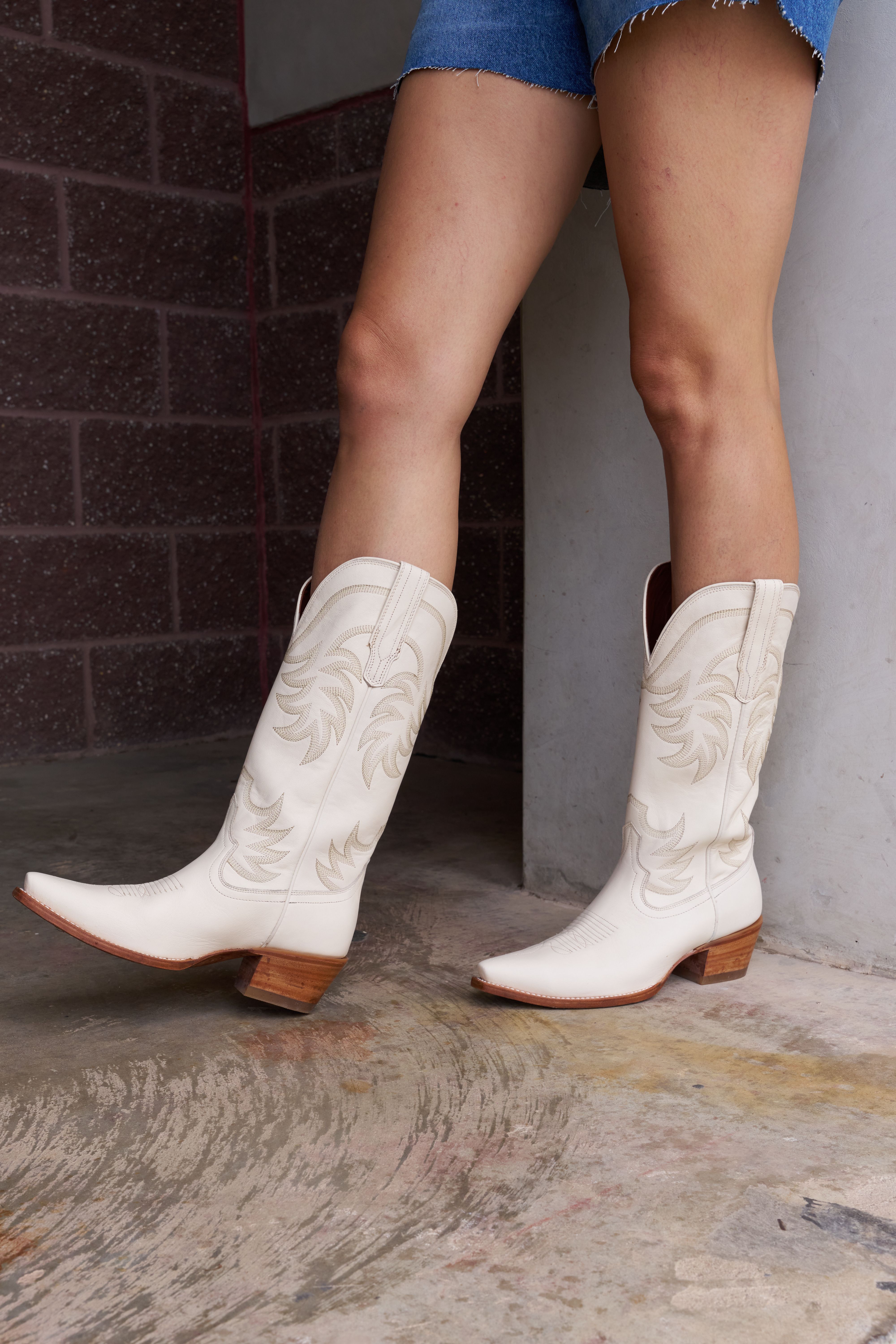 A person stands in white embroidered cowboy boots and frayed denim shorts on a concrete floor near a brick wall.