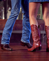 Two individuals dance closely, one wearing blue jeans and brown boots, the other in a blue denim skirt and red cowboy boots, on a wooden floor with a blurred background.