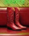 Red cowboy boots with decorative stitching are displayed upright on a red leather bench, with part of a neon "BOOTS" sign visible in the background.