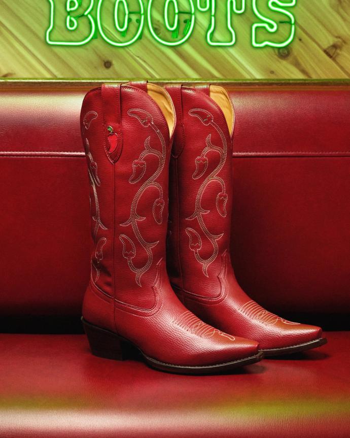 Red cowboy boots with decorative stitching are displayed upright on a red leather bench, with part of a neon "BOOTS" sign visible in the background.