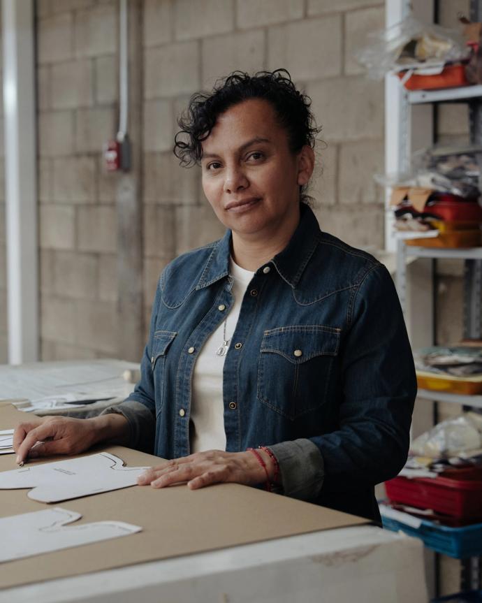 A woman stands at a work table with paper patterns, wearing a denim shirt. Shelves with various items are visible in the background against a brick wall.