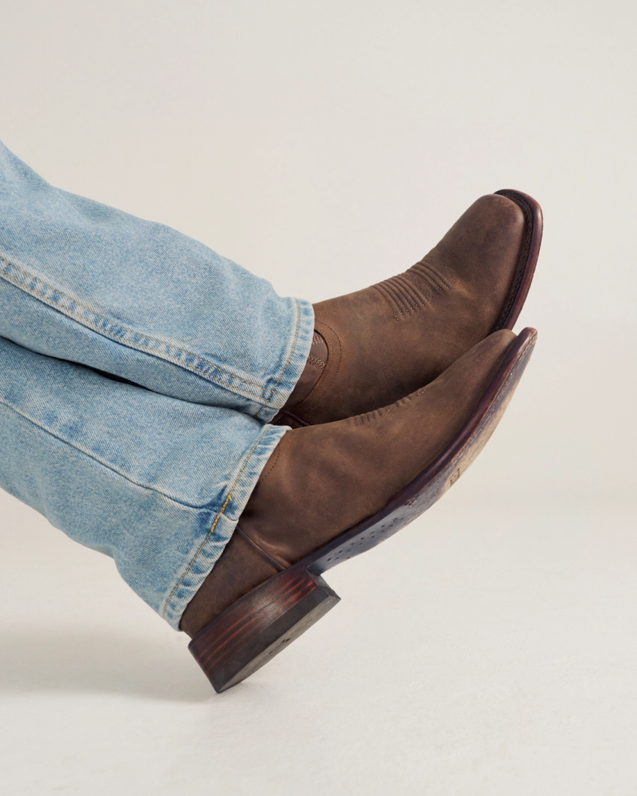 Person wearing light blue jeans and brown leather cowboy boots with legs crossed against a plain background.