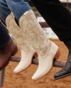 Close-up of three pairs of boots resting on a metal fence. One pair is white with intricate detailing, the other two are brown and black. The ground below is dirt.