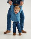 A toddler in a denim shirt and jeans smiles while being held up by an adult, whose hands and legs are visible. Both are wearing brown boots.