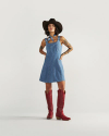 Person in a sleeveless denim dress, black cowboy hat, and tall red cowboy boots stands against a plain white background.