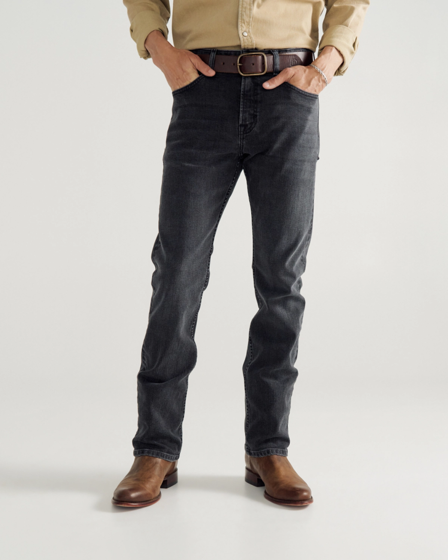 A person wearing a beige shirt, black jeans, a brown belt, and brown leather boots stands against a plain background.