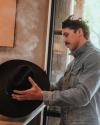 A man in a denim jacket holds a black cowboy hat over rising steam inside a room with wood and glass elements.
