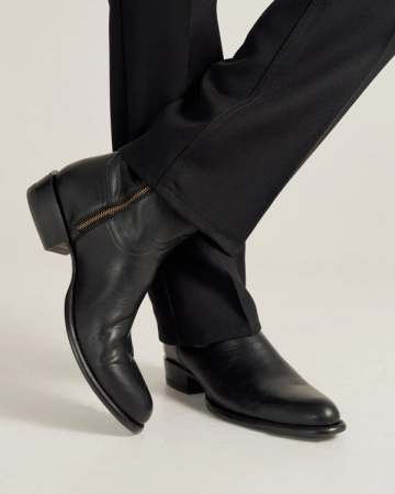 A person wearing black dress pants and black leather boots with a side zipper, standing with one leg crossed over the other.